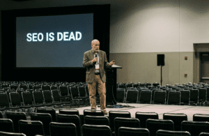 Middle Aged on stage making a presentation called SEO Is Dead to theatre full of empty chairs.